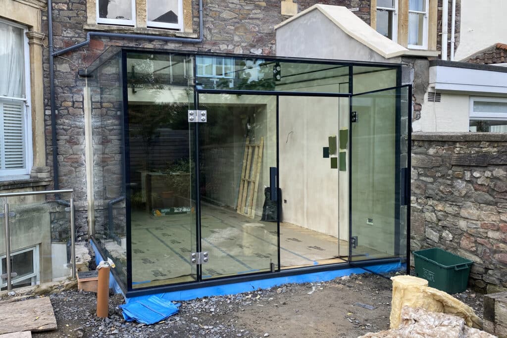 External view of the nearly complete glass kitchen extension under construction in Bristol, featuring clear glass beams and minimal framing.