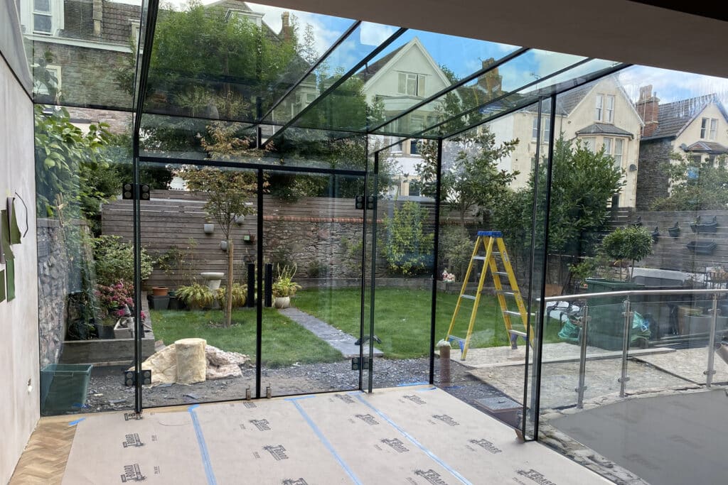 Inside view of the new glass kitchen extension during construction, showing installation of glass beams and frameless glazing panels.